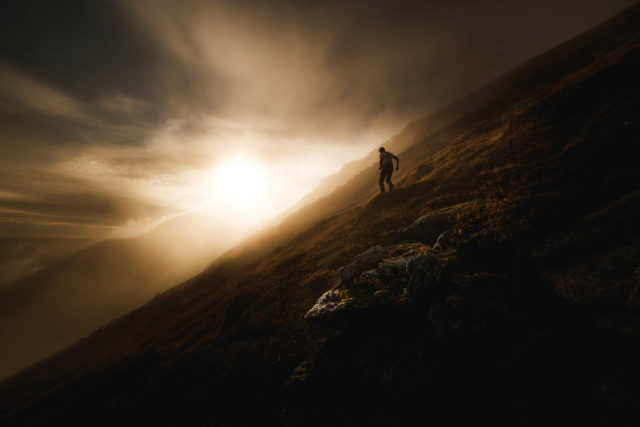 Silhouette sur les pentes des Lofoten Silhouette d’un homme marchant sur une pente herbeuse des Lofoten, éclairée par une lumière rasante de fin de journée